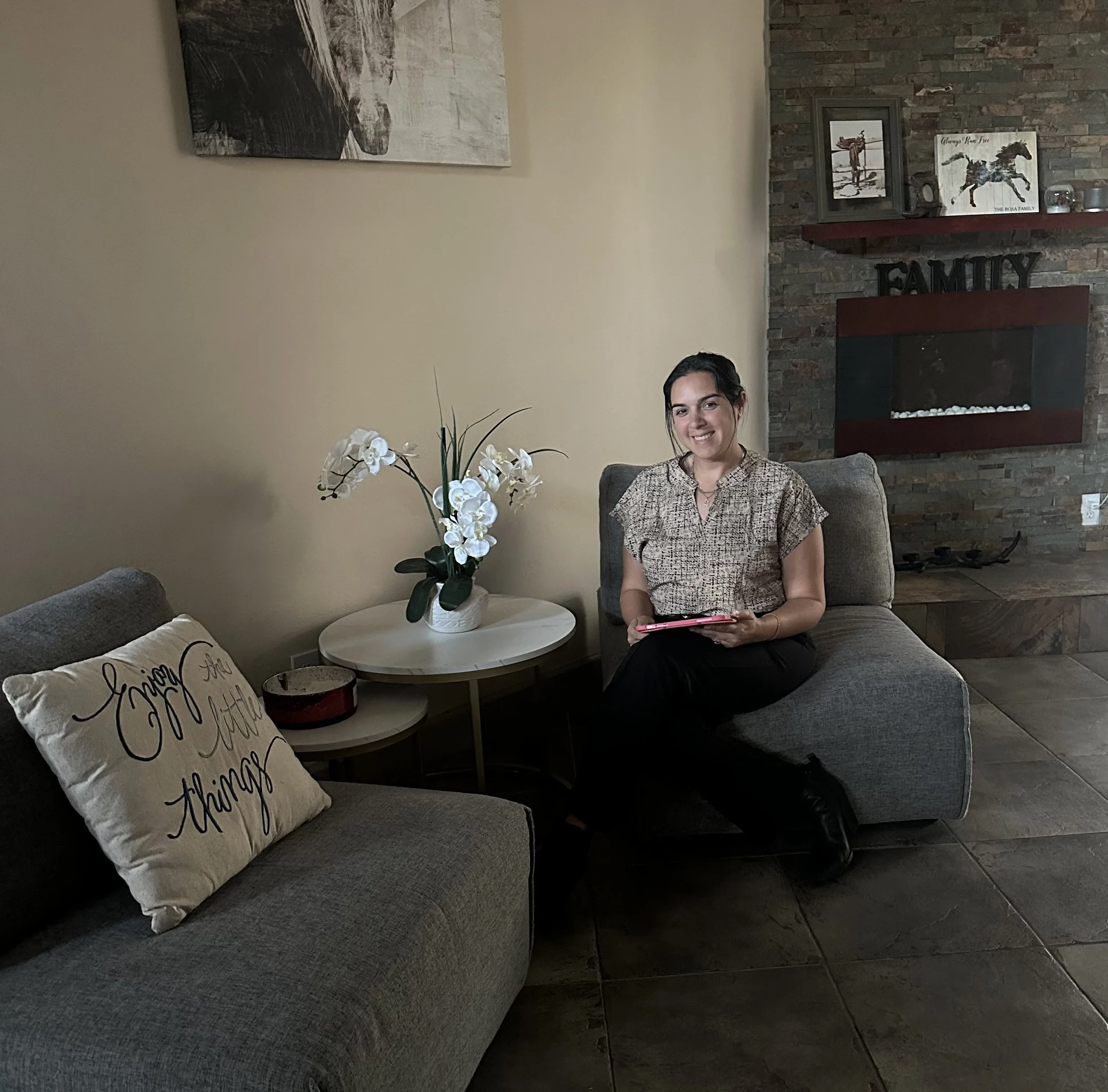 Young woman sitting on a gray sectional sofa in a living room, holding a pink tablet, smiling at the camera. The room features a white side table with a white orchid plant, decorative pillows, and framed artwork on the wall, including a pillow with the words 'Enjoy the little things' and a brick fireplace with family decor.