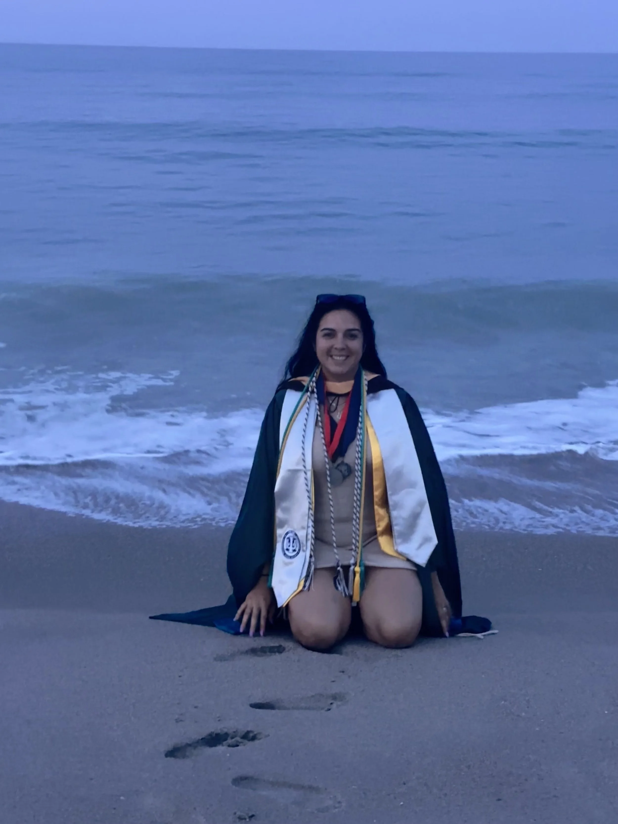 A woman in graduation gown and honor cords kneeling on the beach at sunset, with the ocean in the background.