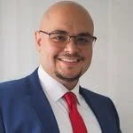 A professional man with a shaved head, glasses, wearing a blue suit, white shirt, and red tie, smiling.