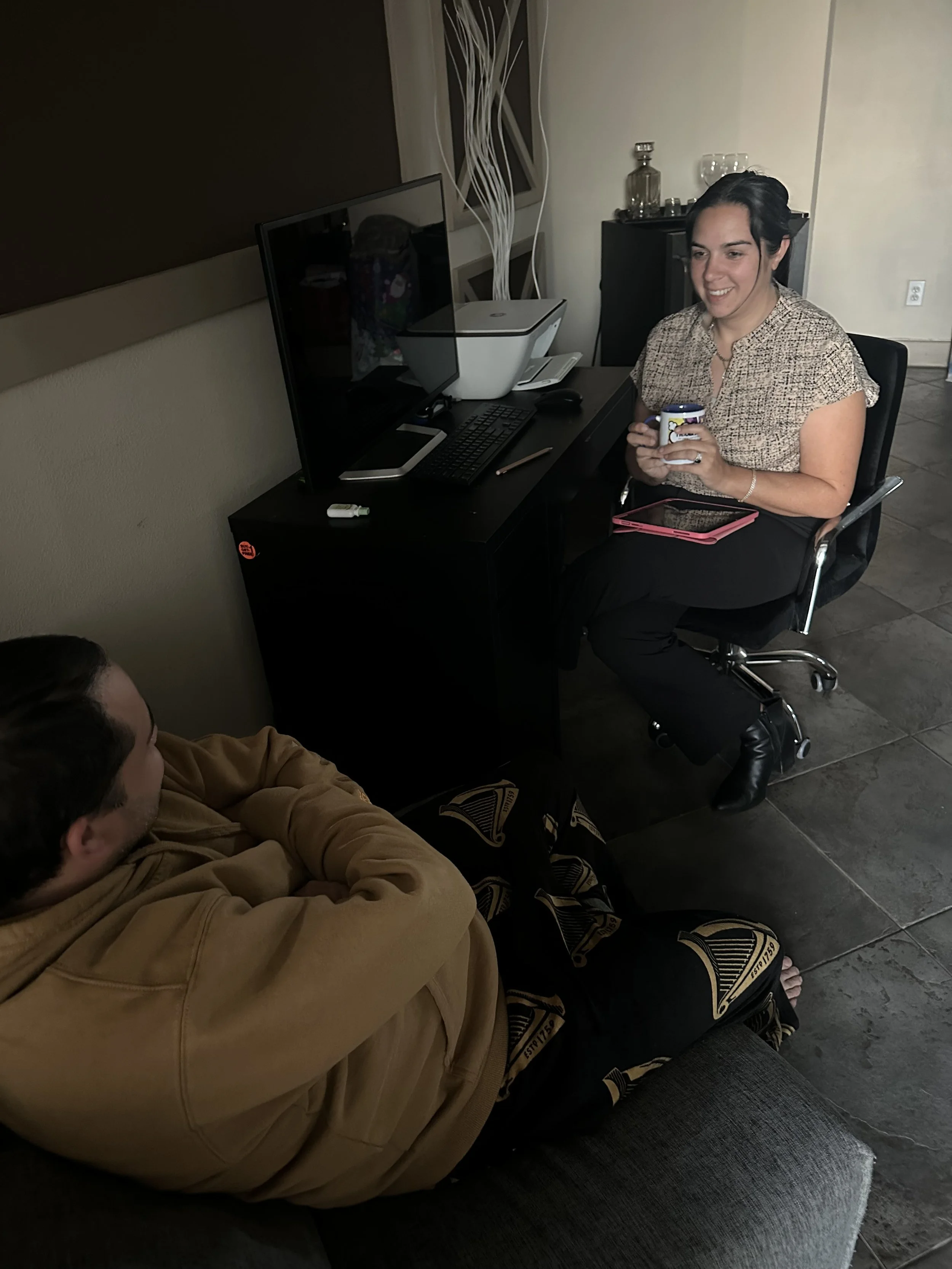 A woman sitting at a desk holding a mug, smiling, and a man sitting on a couch with arms crossed looking at her. The desk has a computer monitor, keyboard, mouse, and a printer. The setting appears to be a home or office with dark tiled flooring.