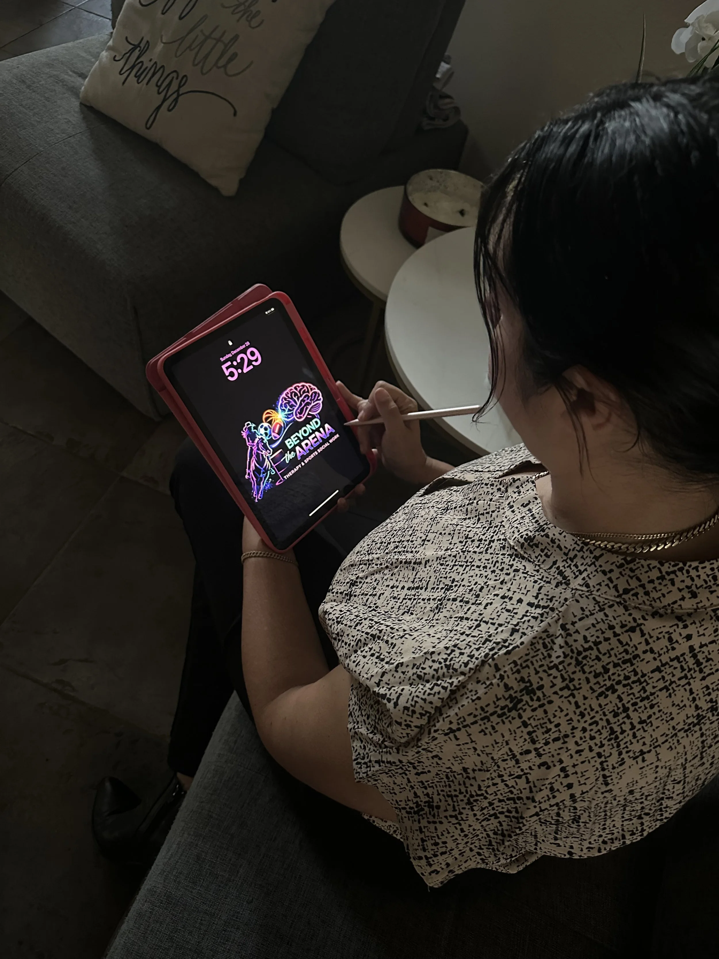 A woman sitting on a gray couch, using a digital pen to draw on a tablet with the screen showing the logo for 'Beyond the Arena' therapy app, in a room with a side table, a pillow with writing, and a decorative flower.
