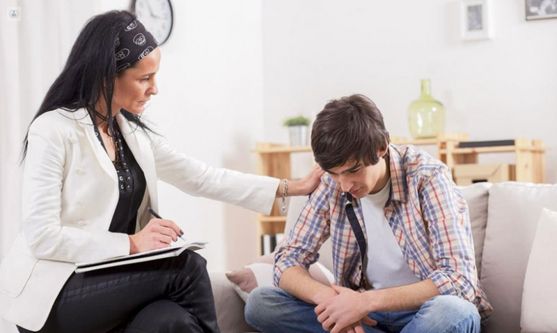 A female therapist is gently comforting a young man who appears distressed, seated on a sofa in a therapy session.