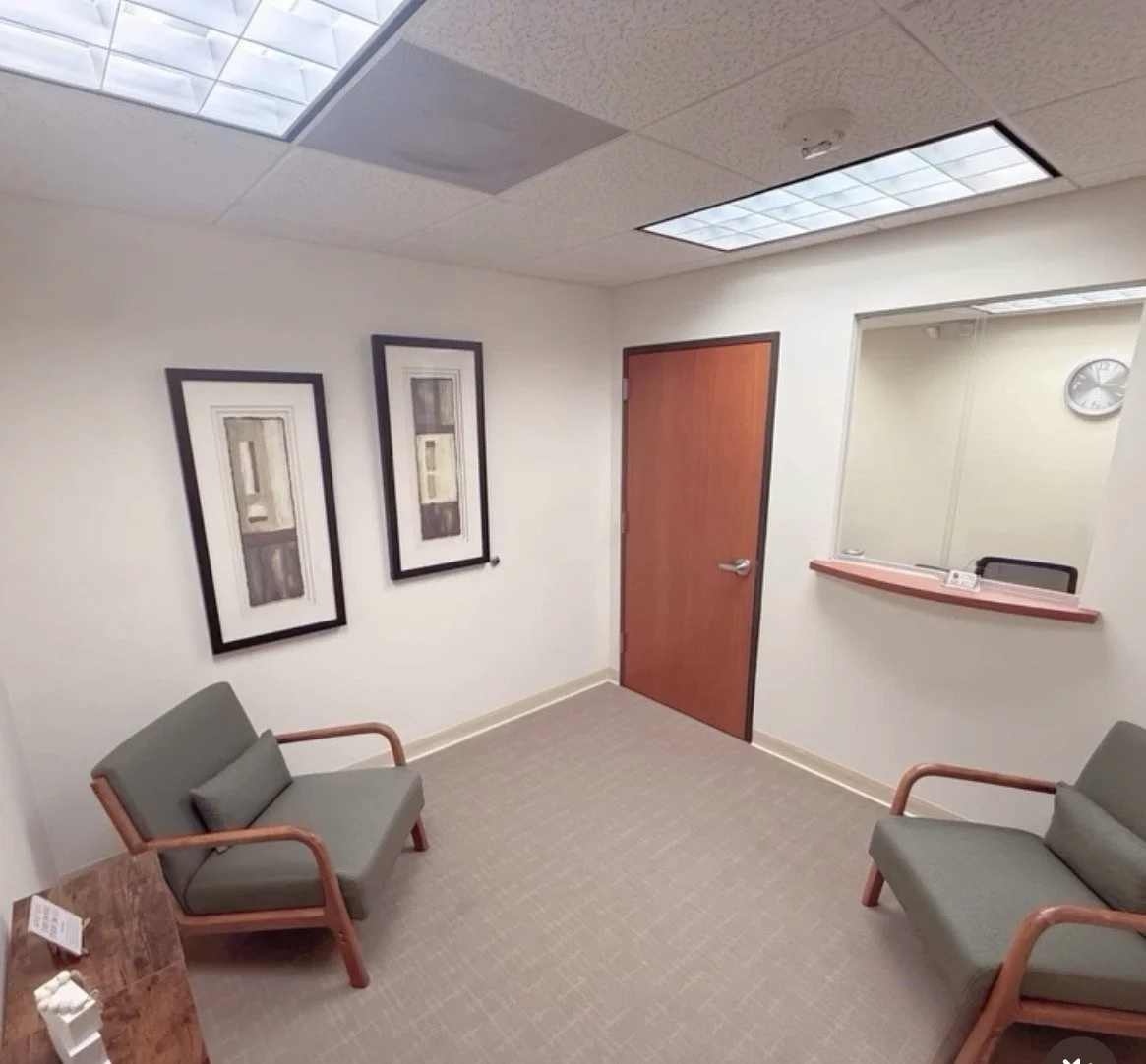 A small waiting room with two green armchairs, a wooden table with tissues, framed abstract art on the wall, a wooden doorway, a window with a counter, and a clock on the wall.