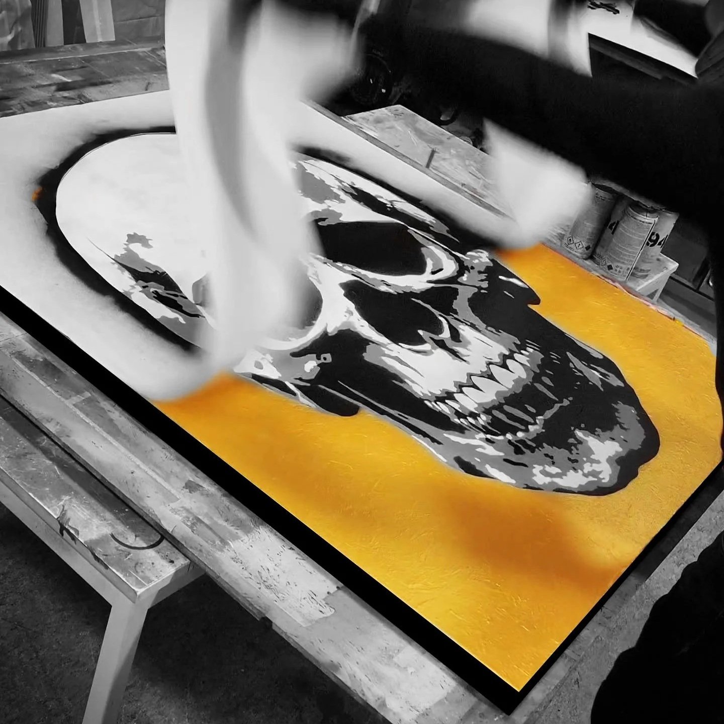 A large, vibrant painting of a human skull with a black and white color scheme on a yellow background, in progress on a worktable.