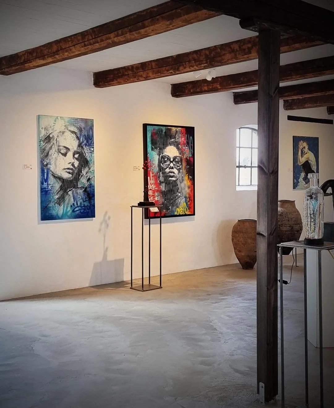 Art gallery with three large portraits of women on white walls, exposed wooden ceiling beams, a small window, decorative vases, and a display stand.