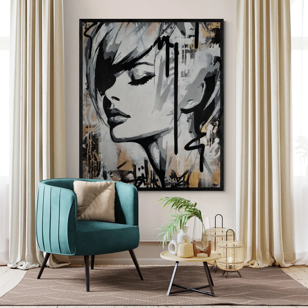 A living room with a large black, white, and gold abstract portrait painting of a woman on the wall. There is a teal armchair with a beige pillow, a small round coffee table with decorative objects and a green plant. Cream curtains frame the scene.