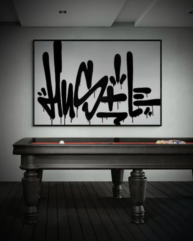 A black pool table with a few balls on it, positioned in front of a white wall with a large graffiti-style artwork, in a modern room with a dark wooden floor.