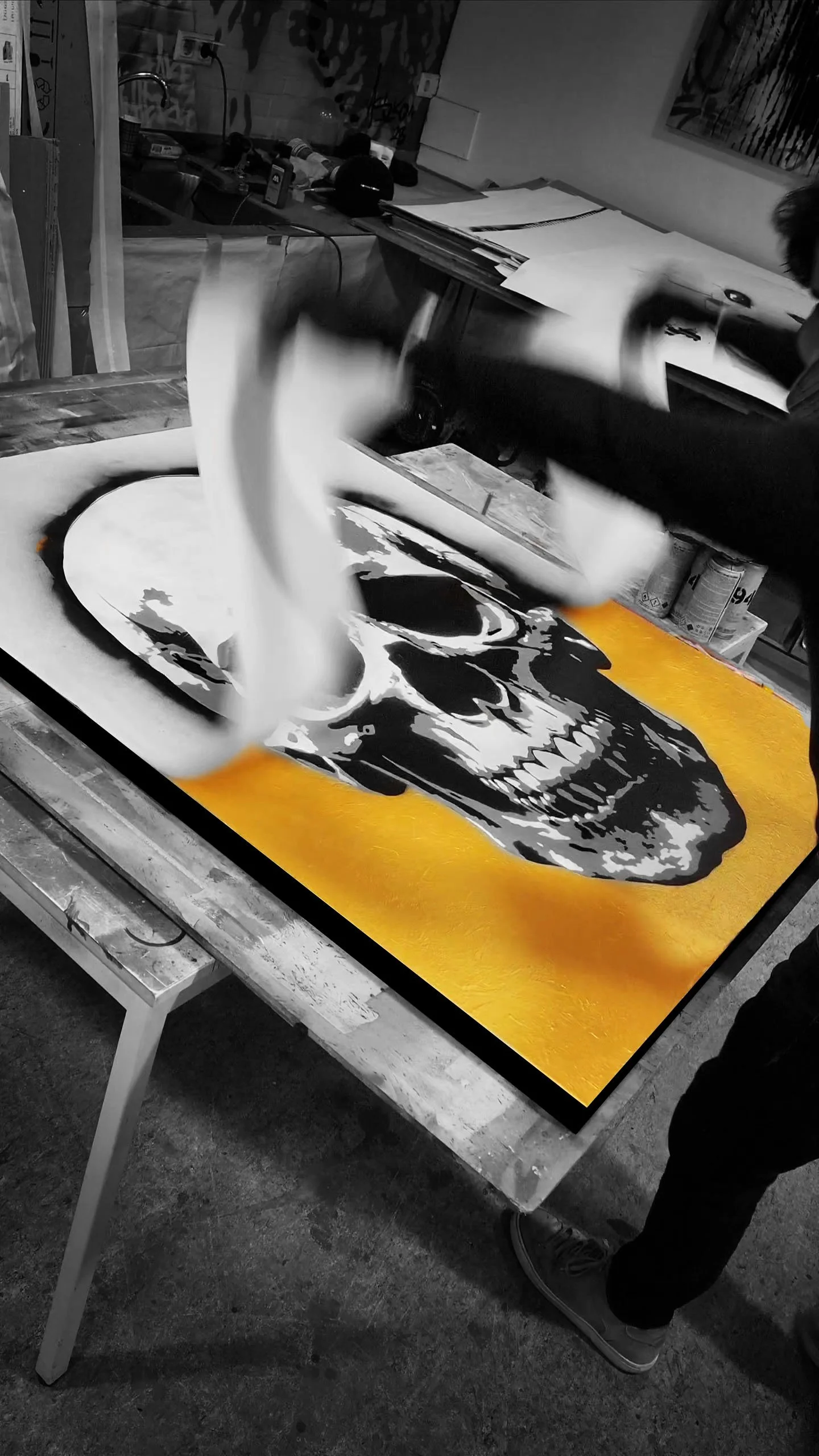 A person is creating a painting of a skull on a canvas, with smoke rising from it. The background shows an art studio with furniture, tools, and supplies.