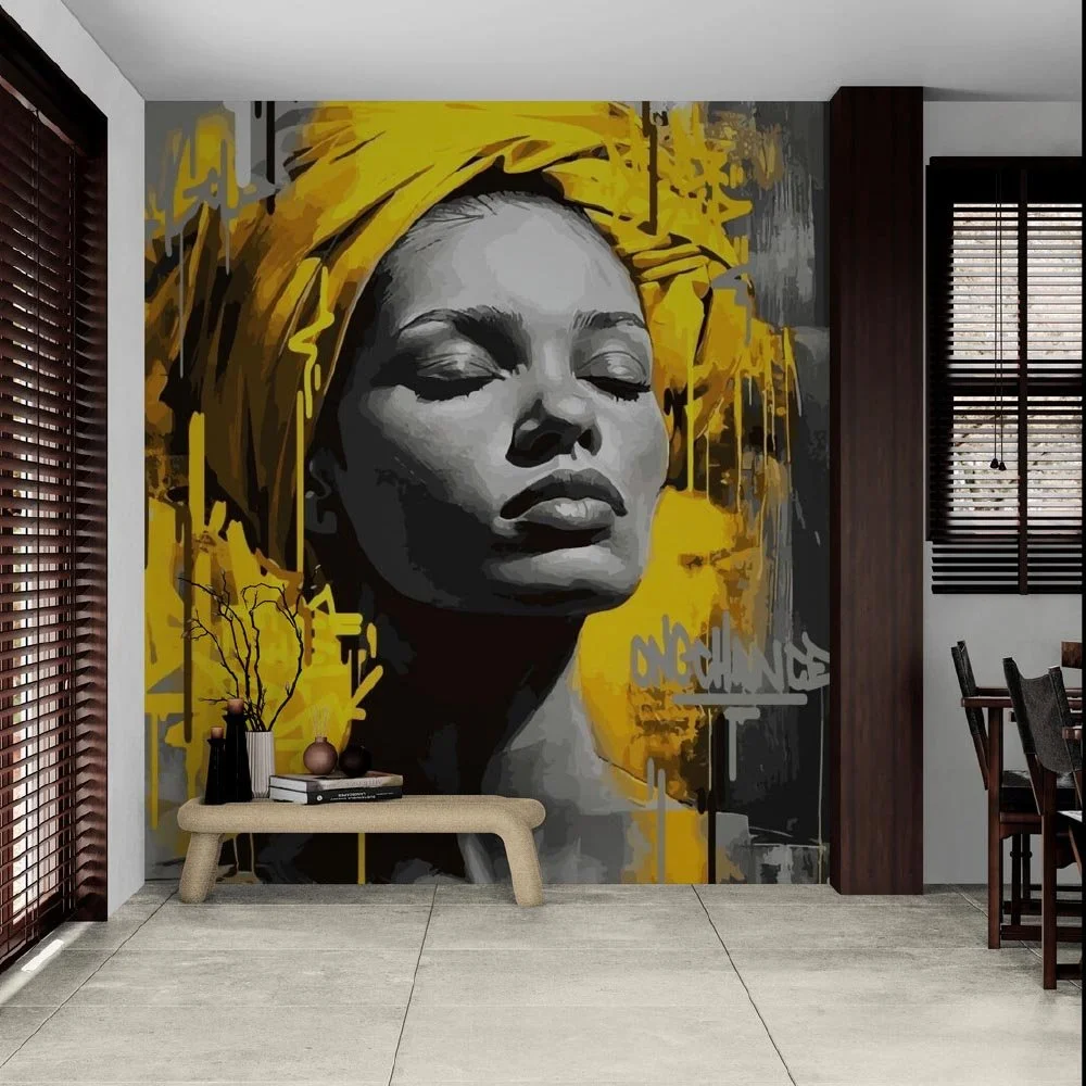 Wall mural of a woman's face with closed eyes, wearing a yellow head wrap, in a modern living room with wooden blinds and a low table with vases.