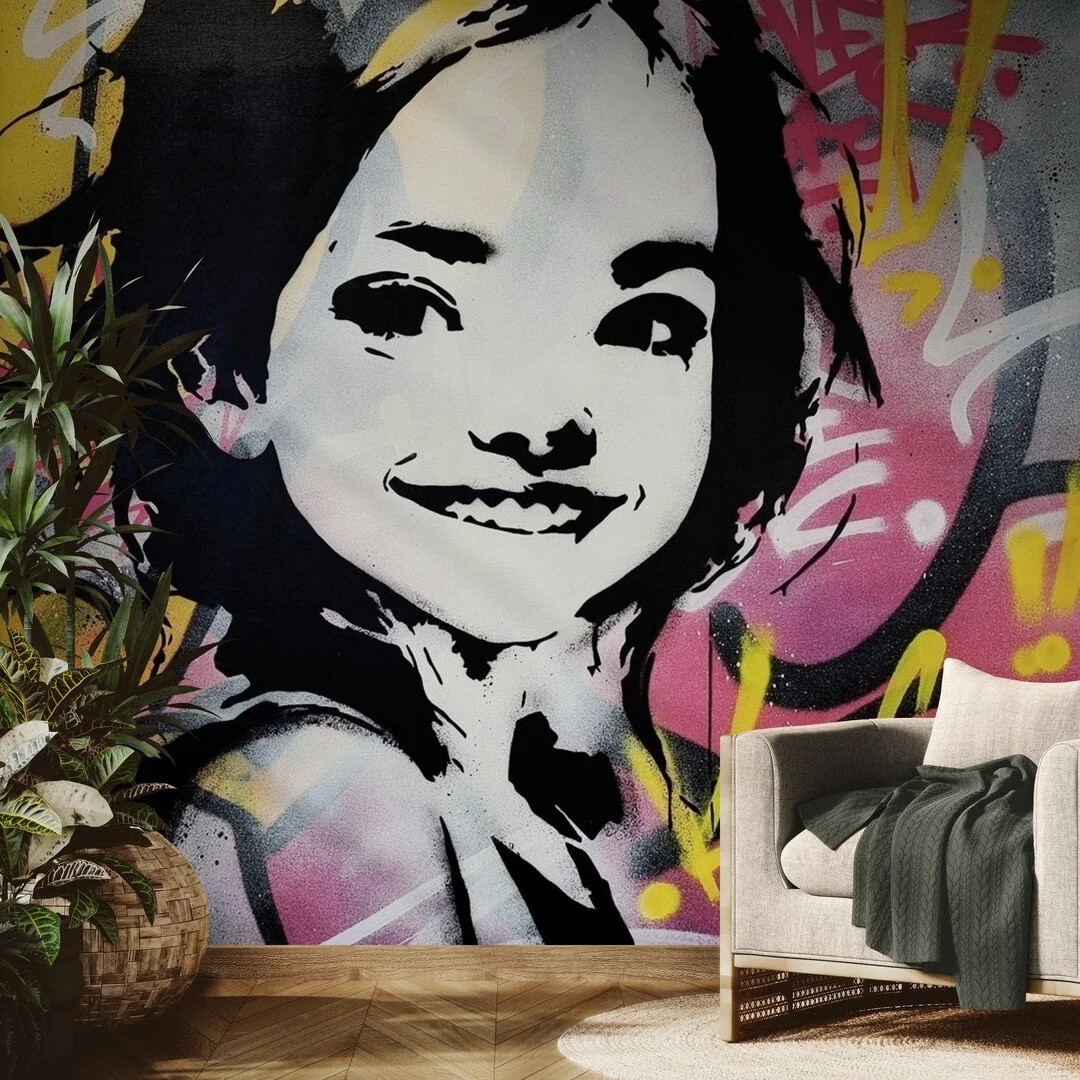 A wall mural of a woman's face in black, white, pink, yellow, and gray graffiti art style, with a plant and a beige couch with a dark blanket in front of it.