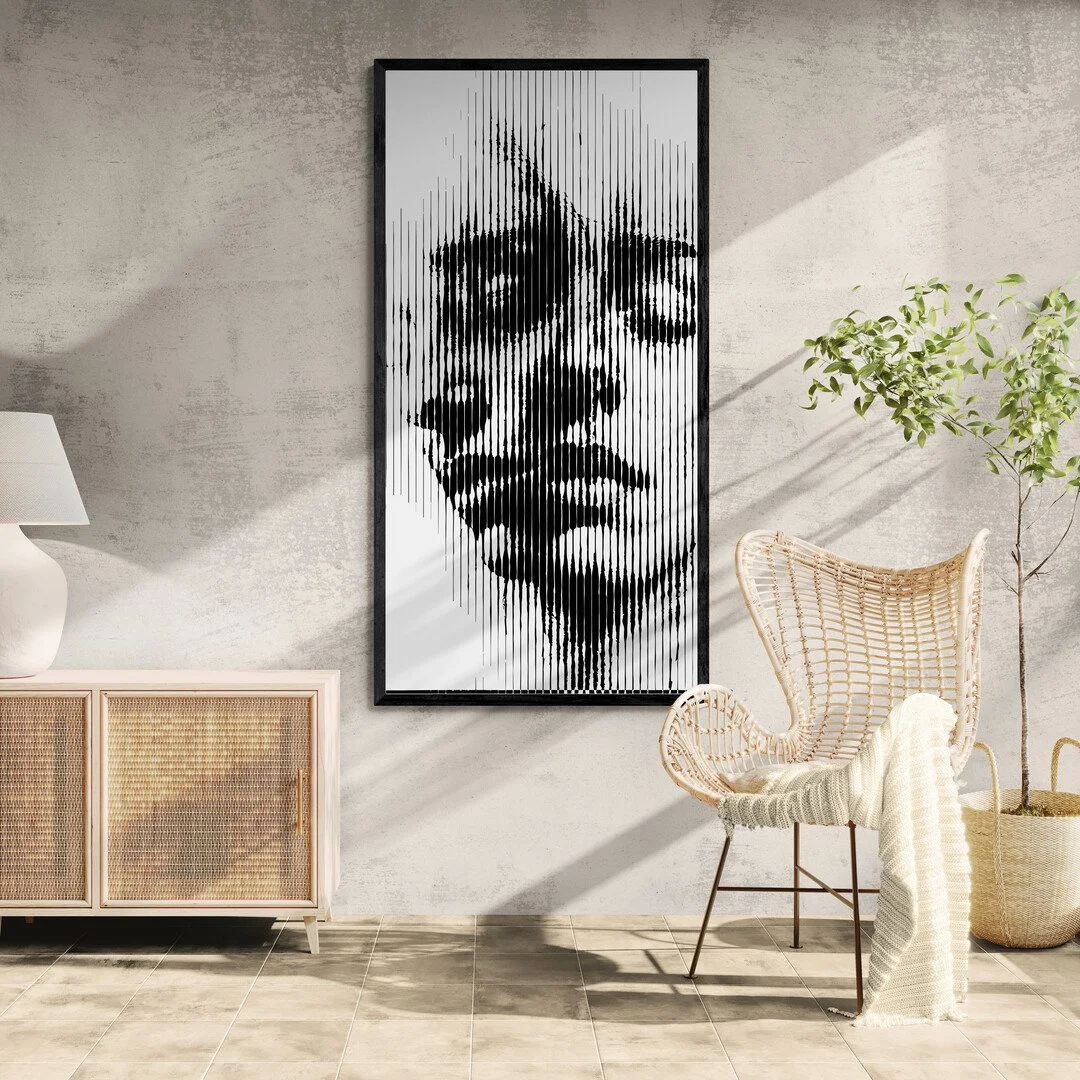 Modern interior with a framed black and white portrait of a woman's face on the wall, a white table lamp, wooden sideboard, wicker chair with a blanket, and a potted plant, with sunlight streaming in.