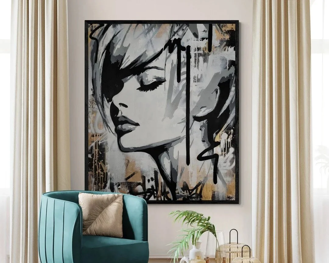 A large black-and-white portrait of a woman with short hair hangs on a wall, with cream-colored curtains on either side. In front of the portrait, there is a teal armchair with a beige pillow and a small brown pillow. A potted plant, glass vases, and decorative items sit on a table below the portrait.