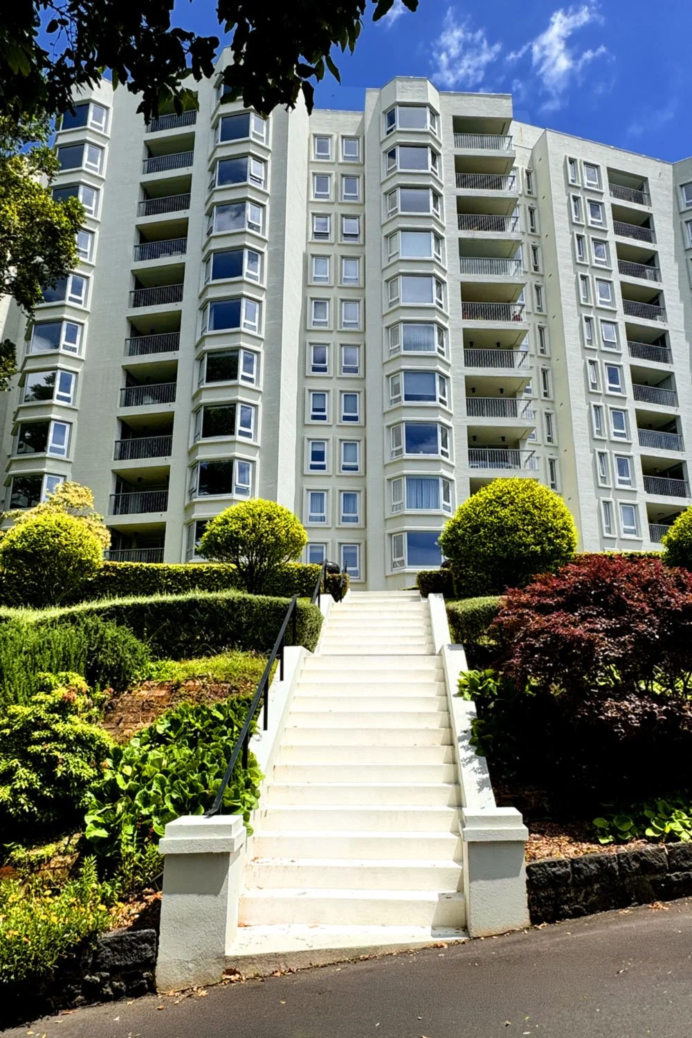 A white high-rise apartment building with multiple balconies and bay windows, surrounded by lush green bushes and trees, with a white staircase leading up to the entrance on a sunny day with blue skies and some clouds.