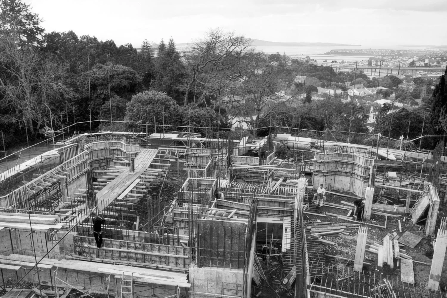 Black and white photo of a construction site on a hillside, with wooden framing, scaffolding, and workers, with trees and a distant landscape in the background.