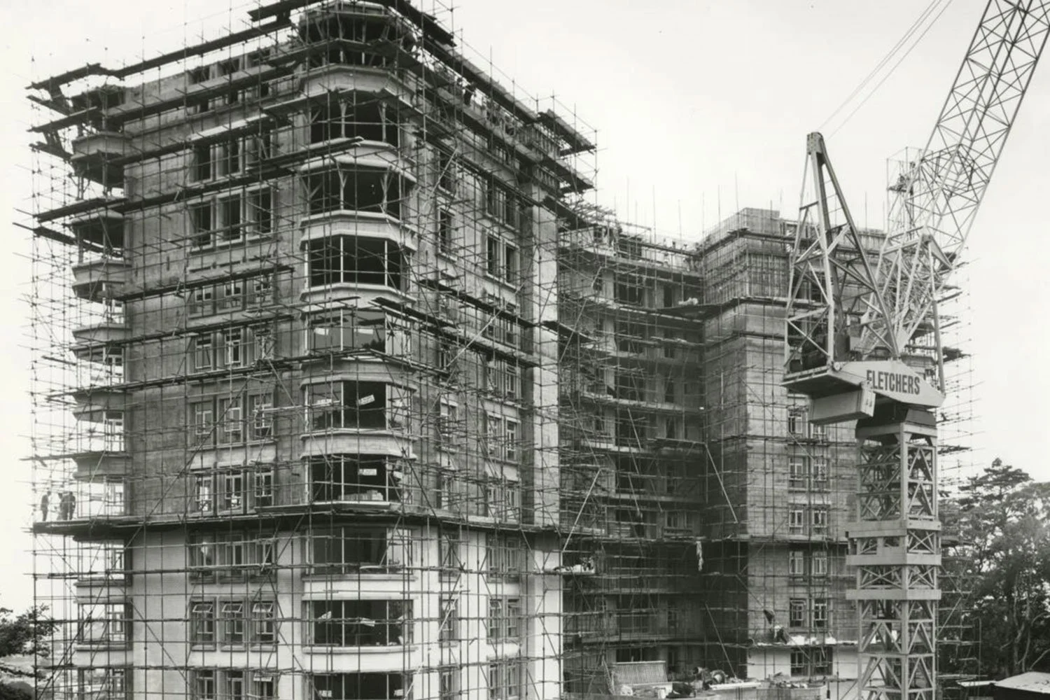 Black and white photo of a multi-story building under construction, surrounded by scaffolding. A crane labeled 'Fletchers' is visible on the right side.
