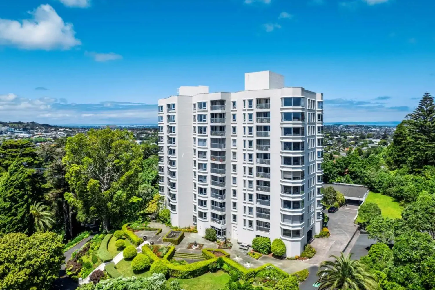The Pines apartment  building viewed from the air, surrounded by lush green trees and manicured gardens, with a parking lot visible in the lower right corner, under a blue sky with scattered clouds.