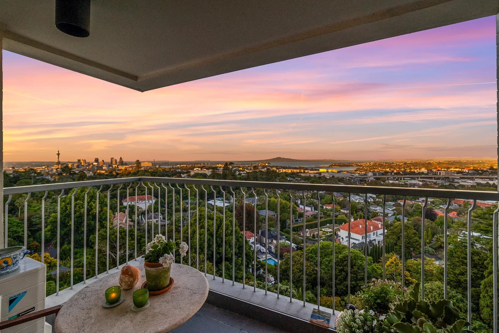 View from the balcony of the Pines apartment showing a cityscape at sunset with colorful sky and a mix of trees, houses, and buildings in the distance.
