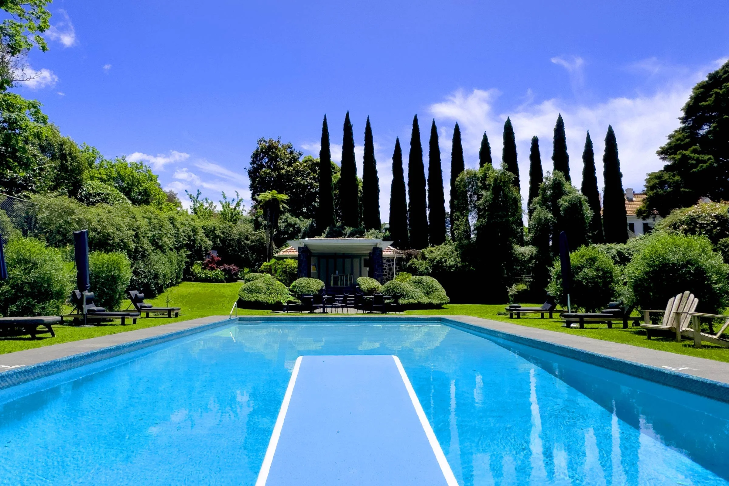 The Pines swimming pool in a landscaped yard with lounge chairs, umbrellas, lush green bushes, and tall cypress trees under a blue sky with a few clouds.