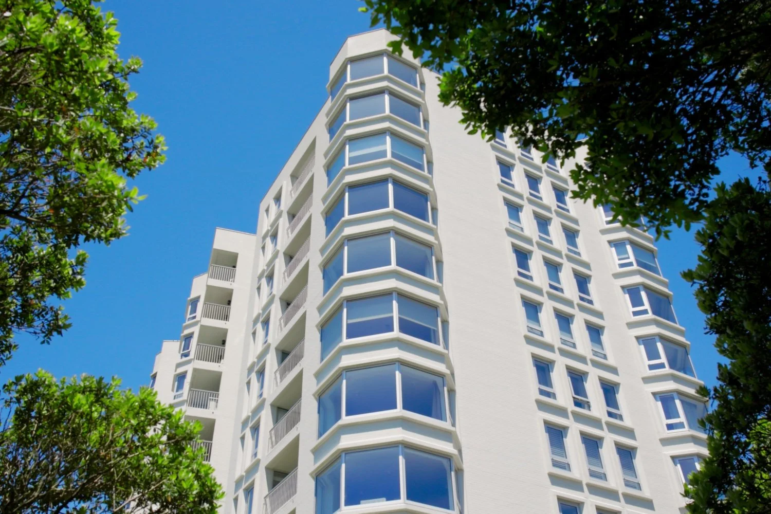 Modernist white residential building with large windows, surrounded by green trees under a clear blue sky.