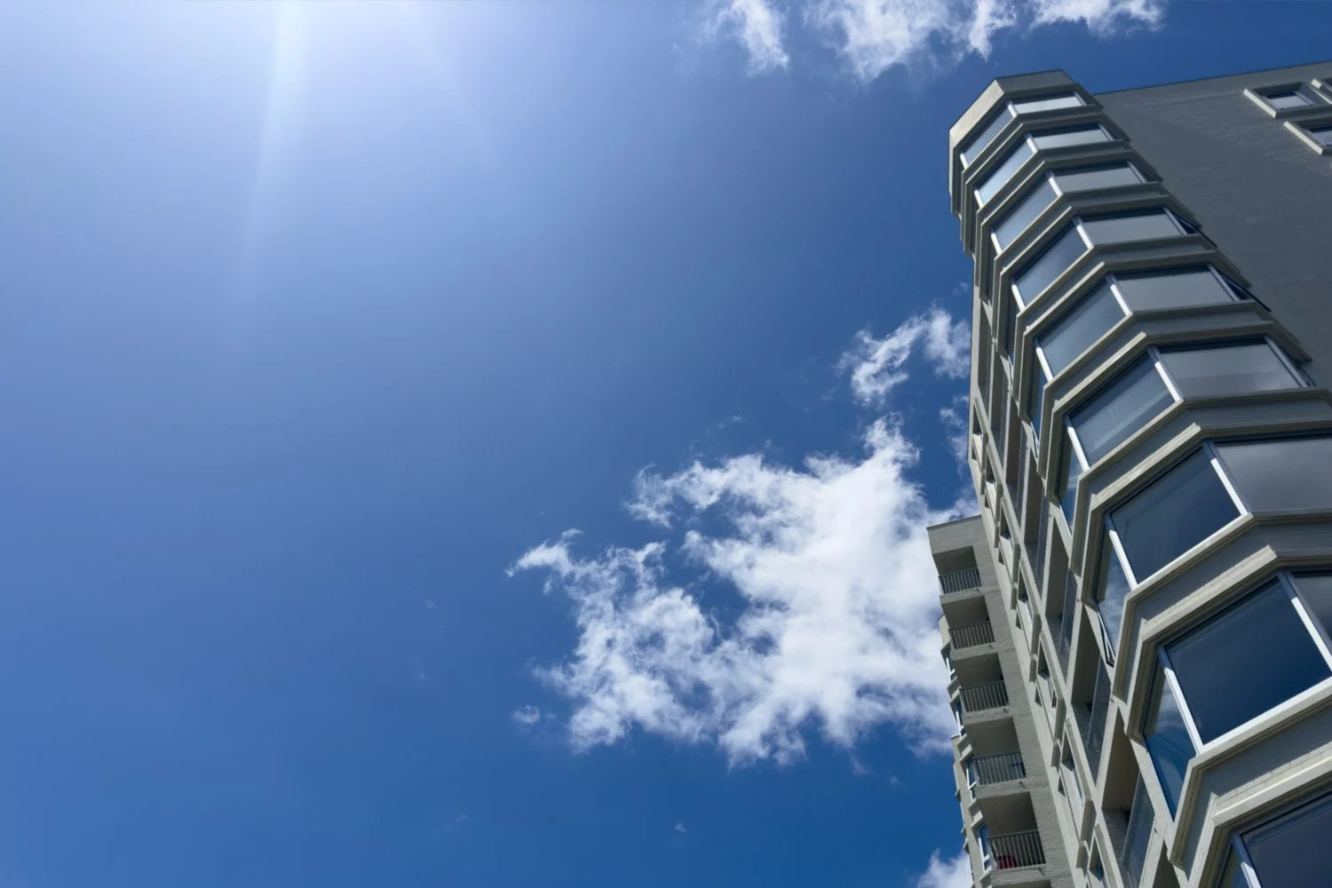 The Pines Apartments , a modernist building with multiple windows set against a bright blue sky with some clouds and sunlight.