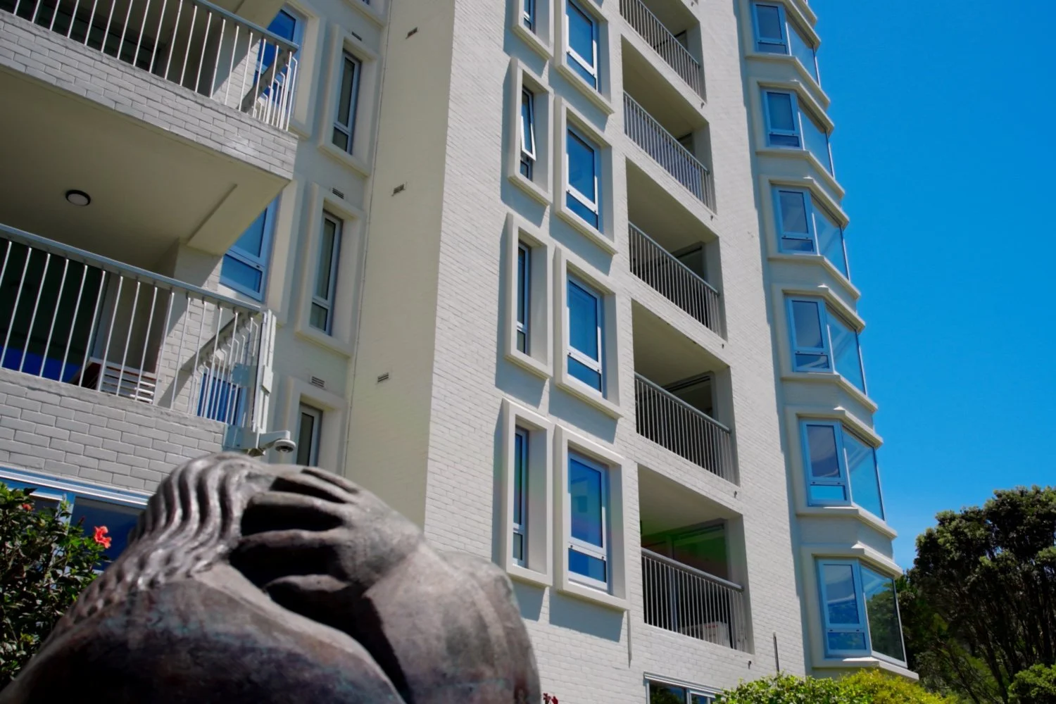 Modernist white multi-story apartment building with balconies and large windows, against a clear blue sky with trees in the background.