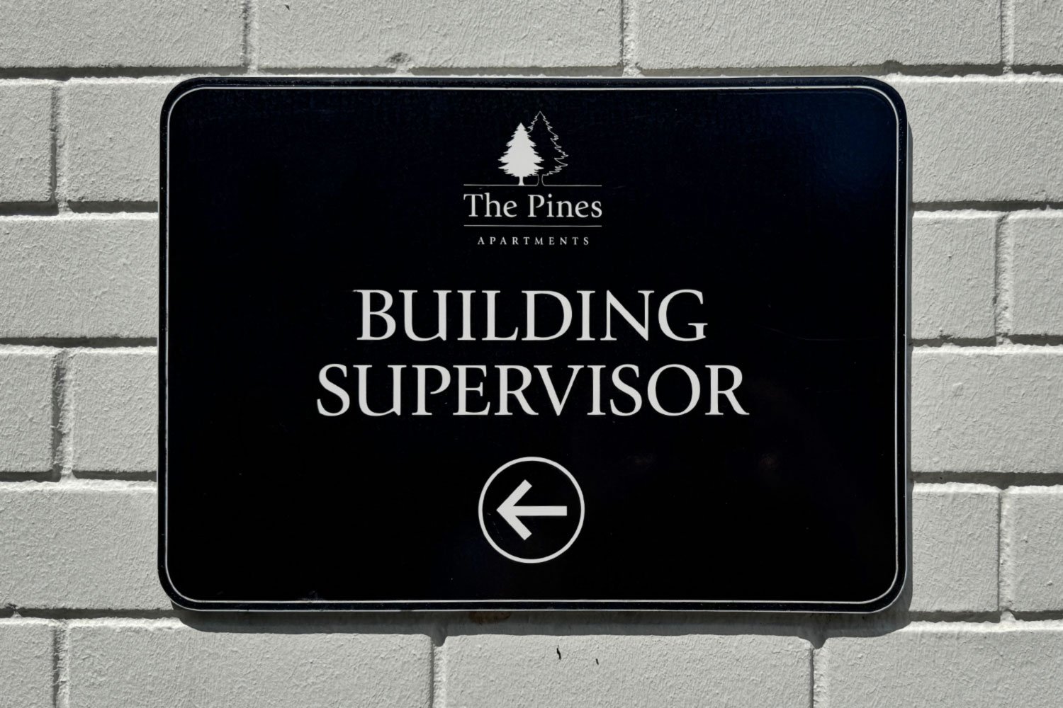 Sign on a white brick wall for The Pines Apartments, indicating the direction to the building supervisor with a left arrow.
