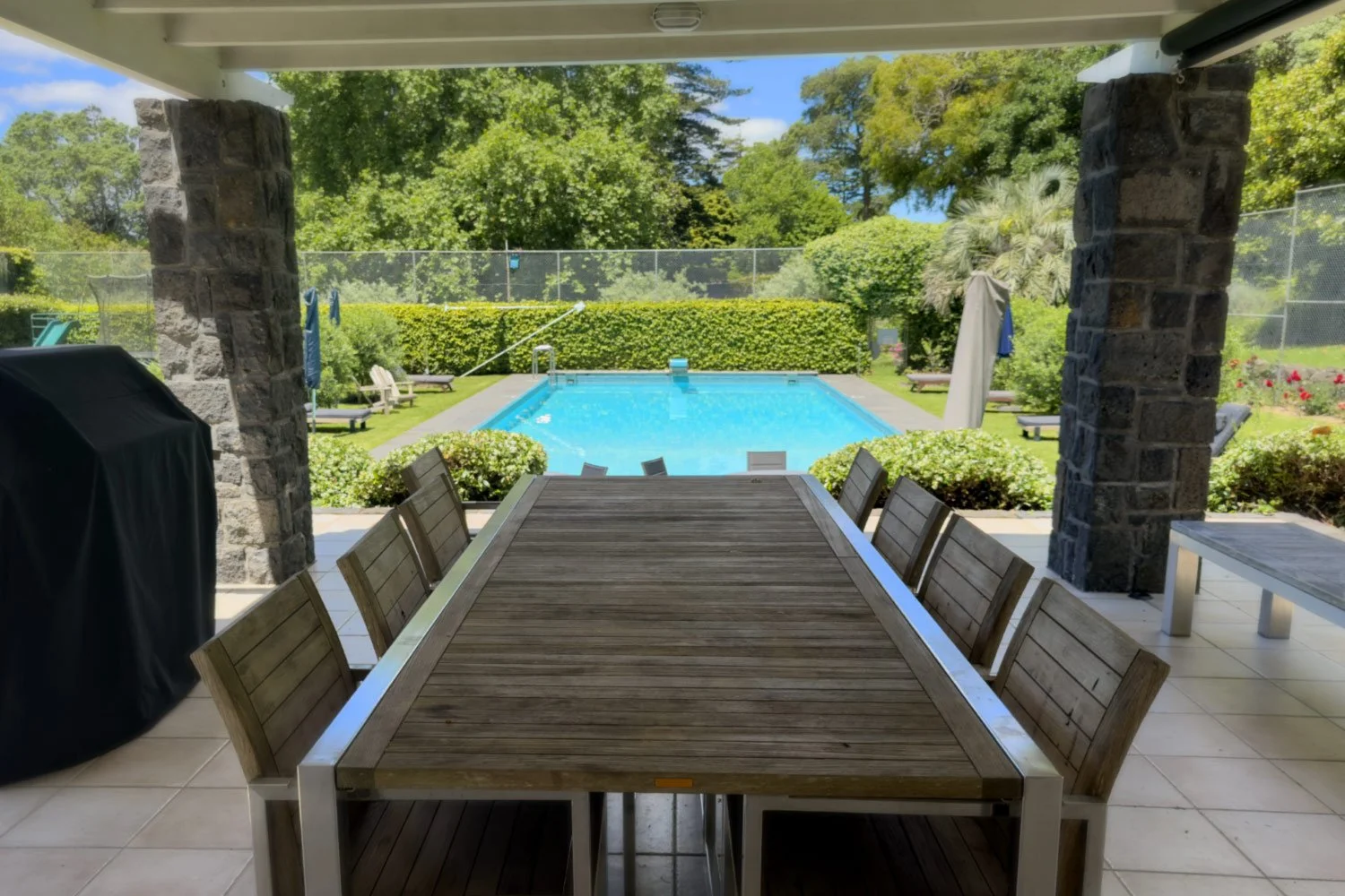 Outdoor backyard pool area with a wooden dining table and chairs under a covered patio, surrounded by green bushes and trees.