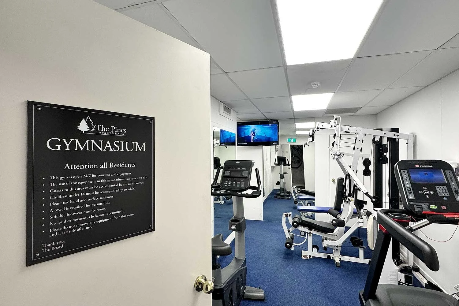 View of a gym room with exercise equipment, two wall-mounted TVs, and a sign on the door listing gym rules at The Pines Apartments.