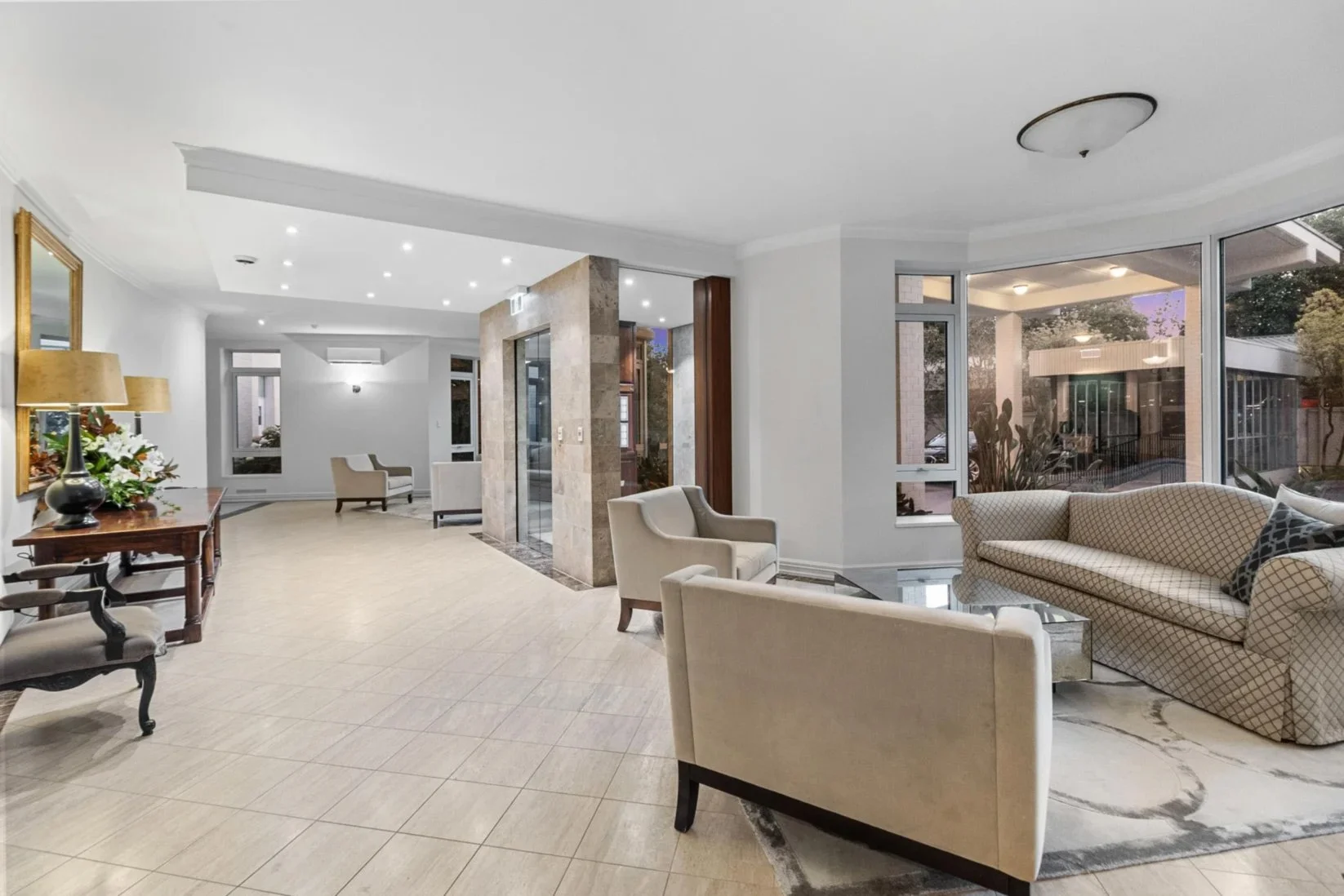 The elegant lobby with white walls, beige tiled floor, large windows, and contemporary furniture including sofas, armchairs, and a wooden table with a lamp and floral arrangement.