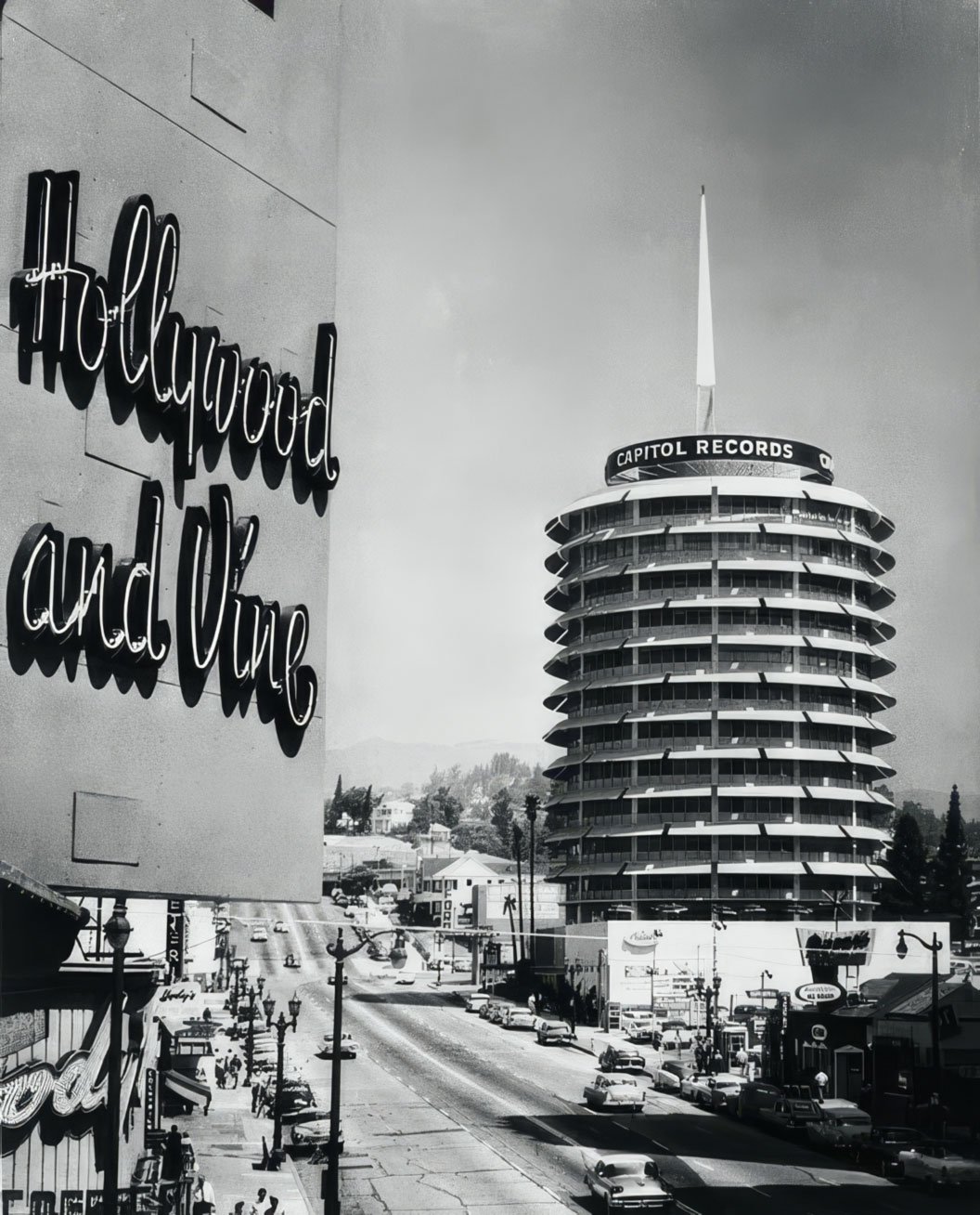 Black and white photo of the Capitol Records building in Hollywood, California, with a sign on a building in the foreground that reads "Hollywood and Vine."