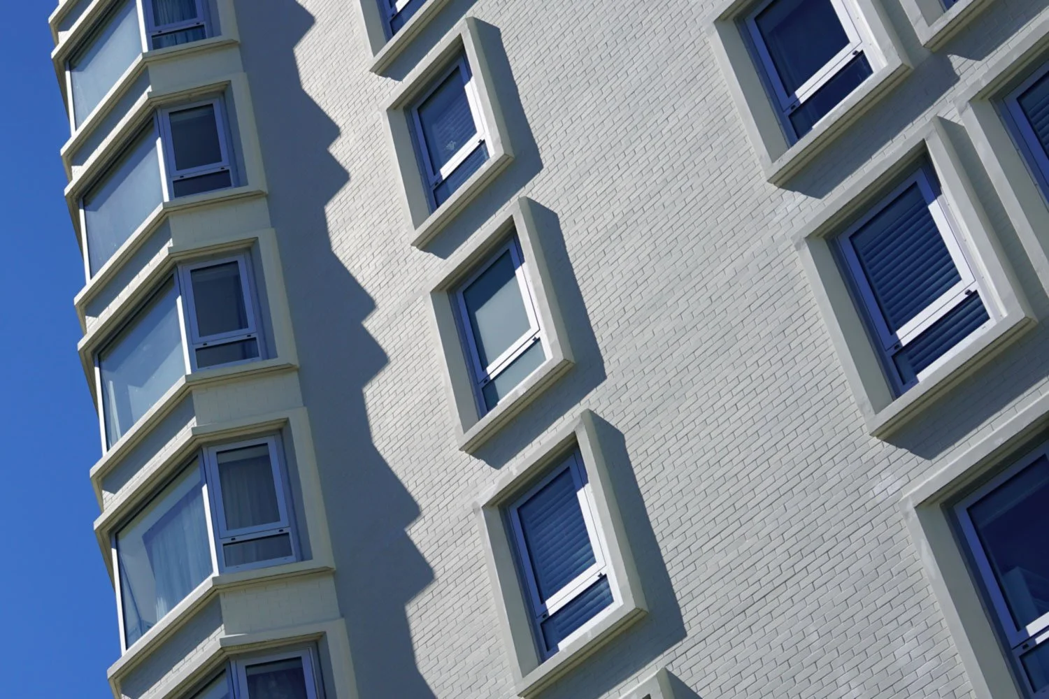 Close-up of a modernist white brick apartment building with multiple rectangular windows, some with blinds, against a clear blue sky. The building's corner protrudes outward, creating shadows.