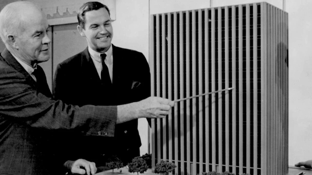 Two men in suits pointing at a large architectural model of a skyscraper.