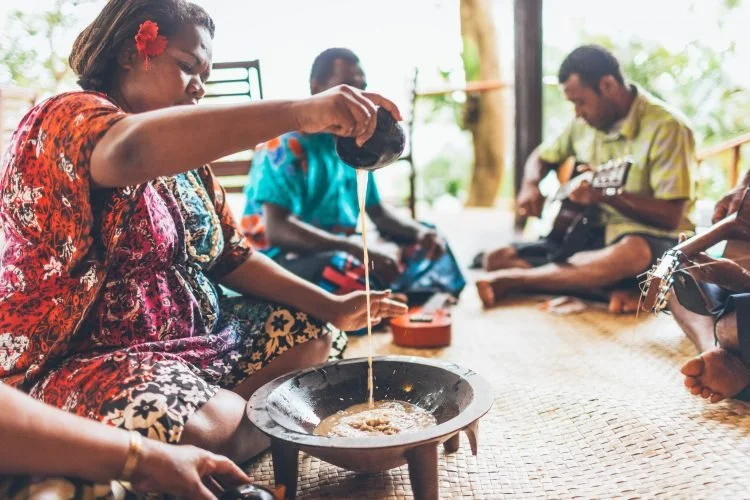 People sitting on a mat, playing guitars, and sharing kava together outdoors under a shaded area.