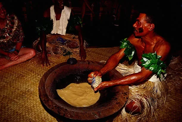 A man with tribal attire, including leaves on his arms, is holding a container over a large clay pot with kava and a small patterned object. Two women and a child sit nearby on a woven mat in a traditional setting.