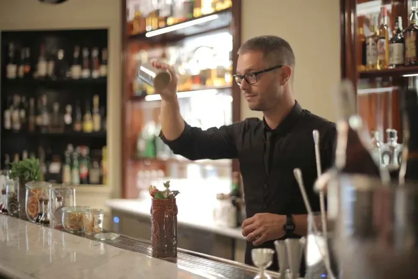 A bartender with glasses and short hair, wearing a black shirt, preparing a drink in a bar with bottles on shelves behind him.