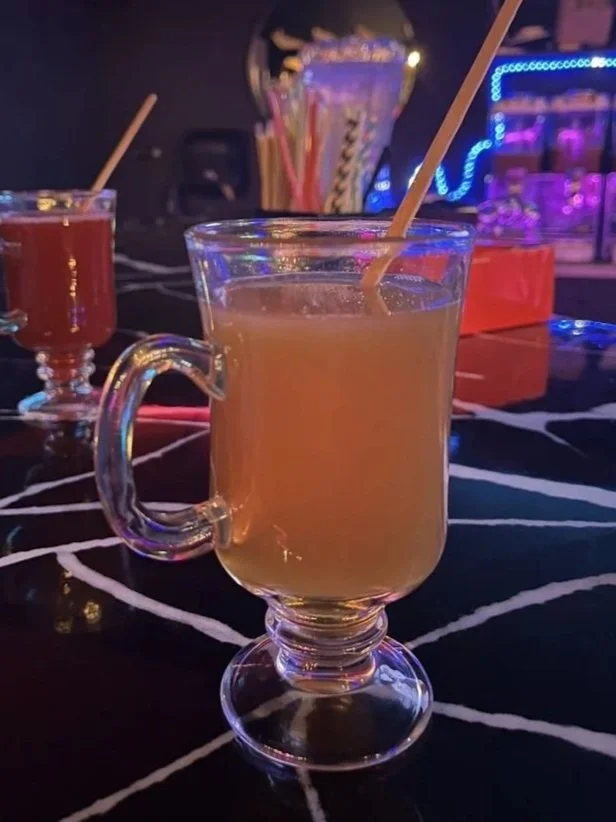 A glass mug filled with kratom, placed on a black table with white lines. In the background, there are other similarly styled drinks and colorful straws, with colorful lights and a bar setting.
