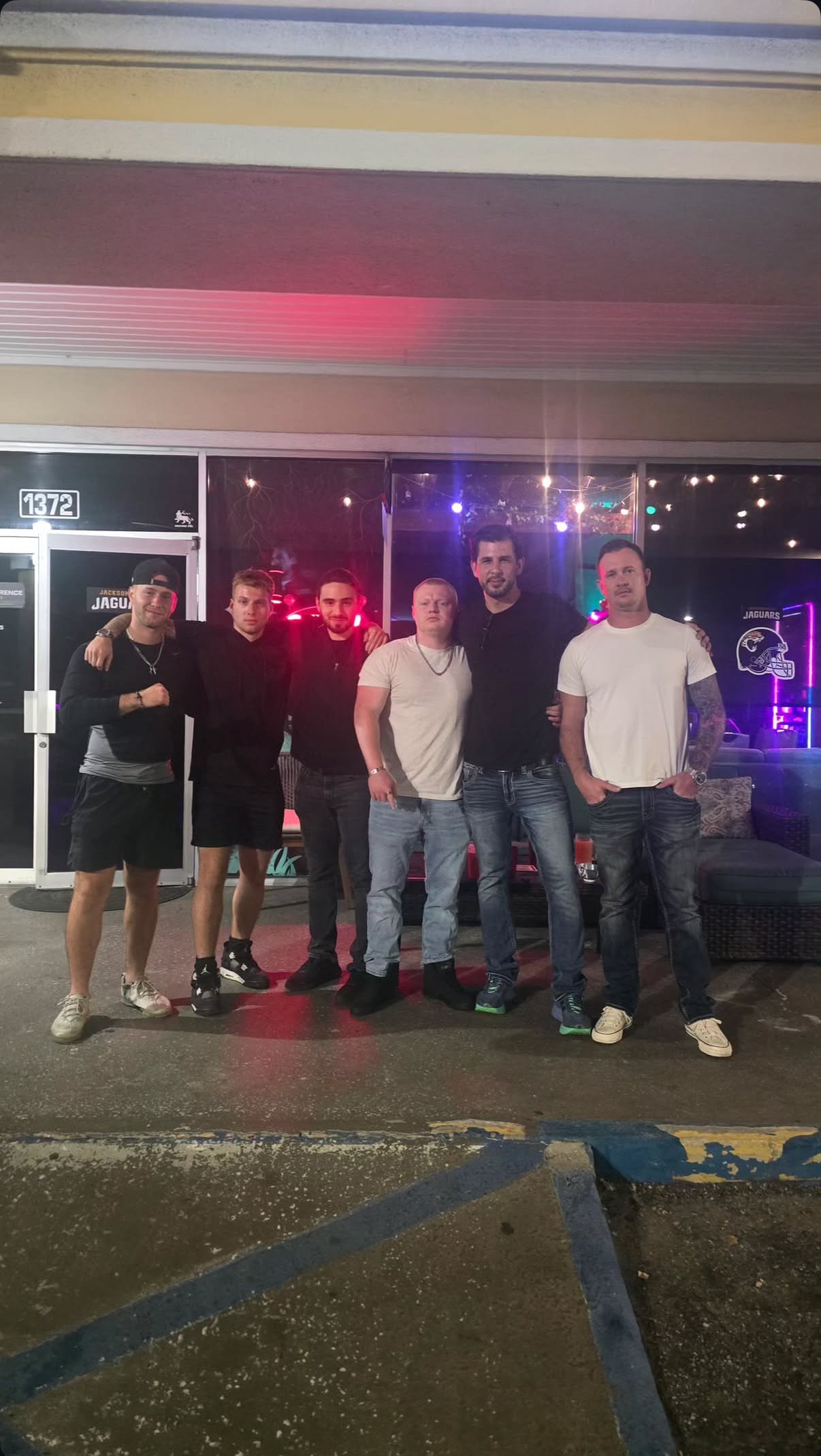 Six men standing in front of a glass door at night, with neon lights and reflections visible in the background.