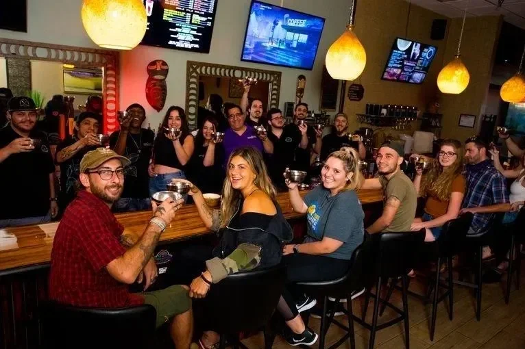 Group of friends at a bar raising glasses, smiling, with a bartender behind the counter, TVs on the wall, warm lighting, and casual atmosphere.