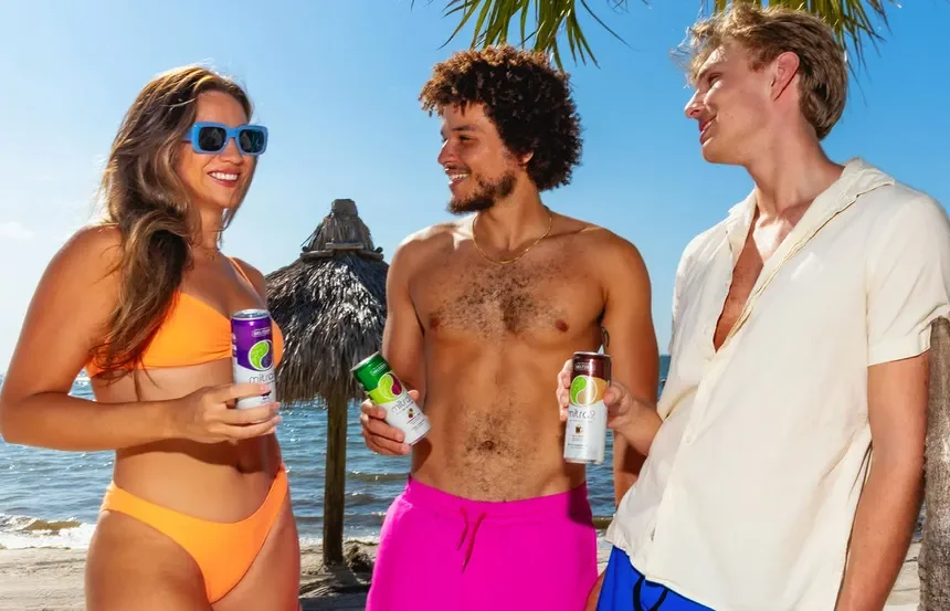 Three young adults at the beach, smiling and holding kratom seltzers, with a thatched umbrella and the ocean in the background.