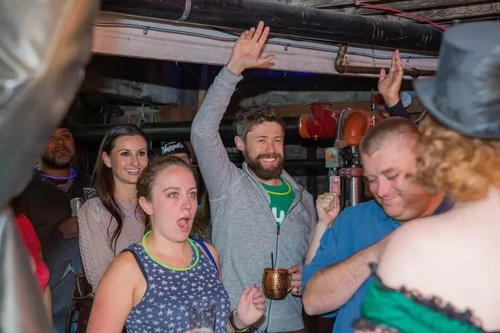 A group of friends celebrating indoors, with one man raising his hand, smiling, and holding a mug, while others around him are smiling or reacting joyfully.
