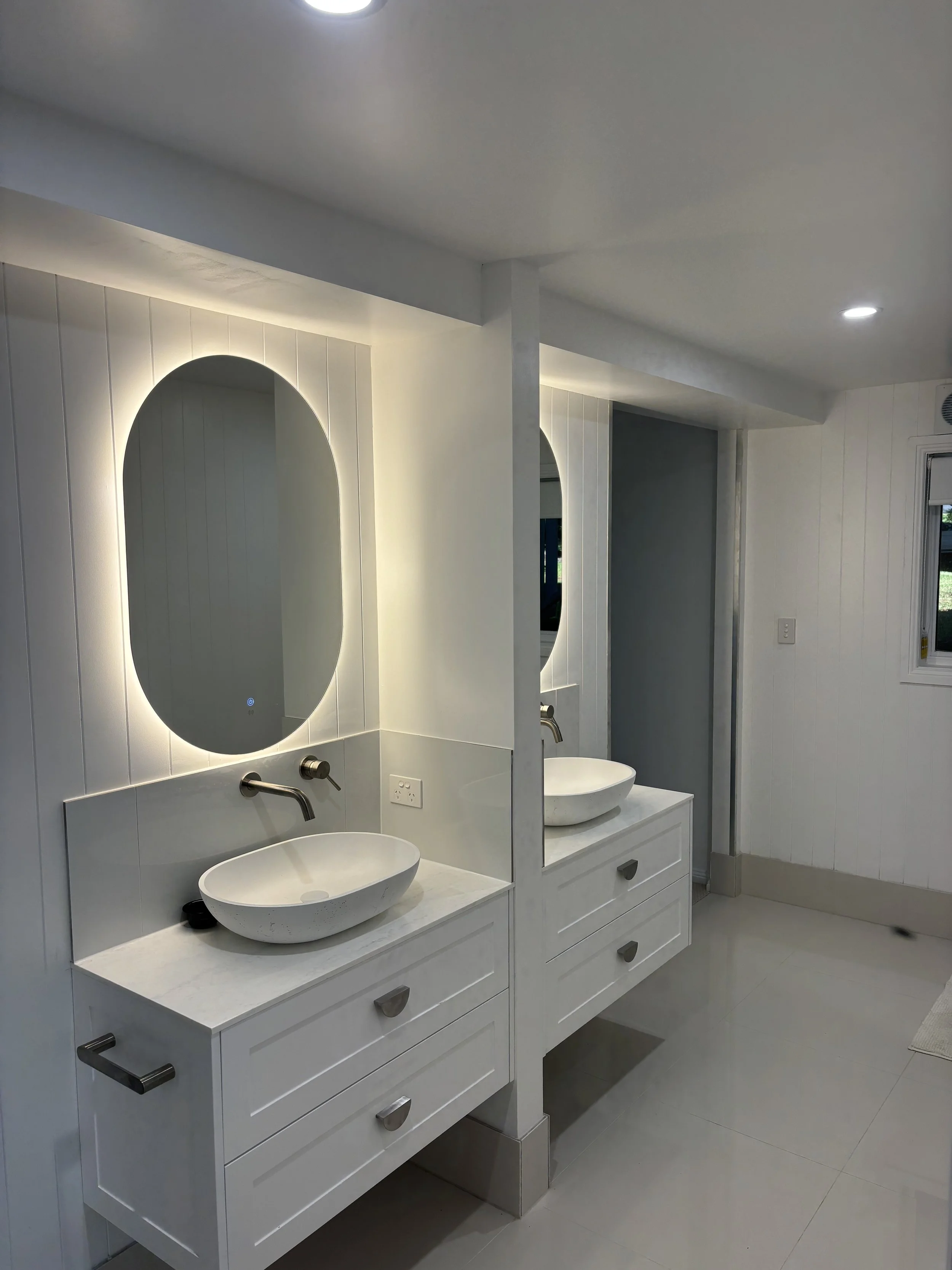 Modern bathroom with two white vessel sinks on white vanities, large oval backlit mirrors, and white tiled walls.