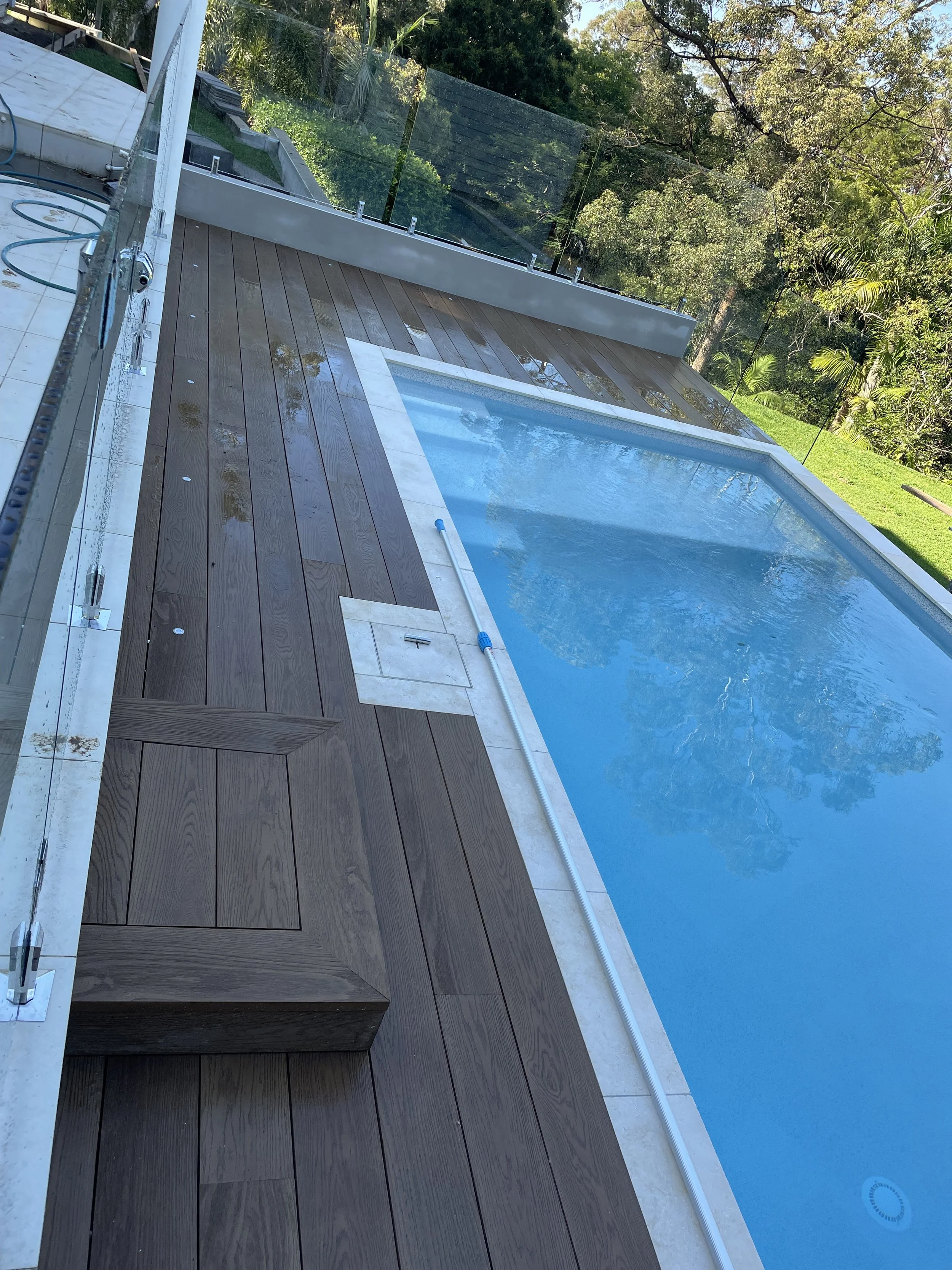 A rectangular swimming pool with blue water, surrounded by beige tiles and a wooden deck, enclosed by clear glass fencing, with trees and green foliage in the background.