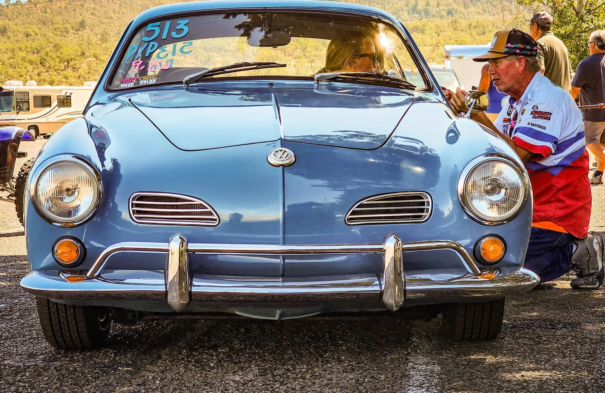 Front view of a vintage blue Volkswagen Karmann Ghia at a car show, with people around and a scenic background.
