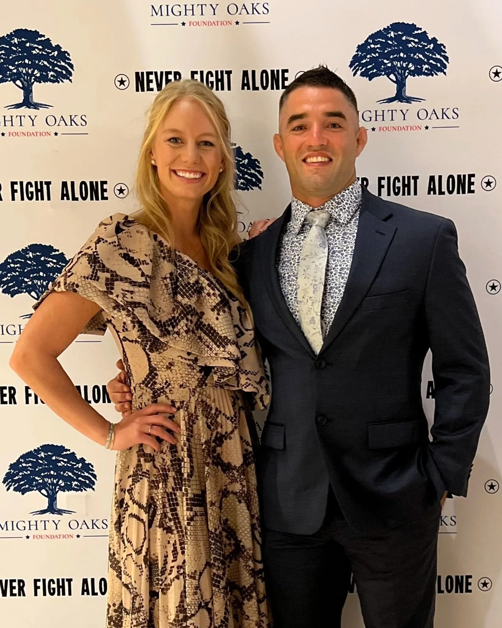A woman and a man dressed in formal attire, standing in front of a backdrop with logos of Mighty Oaks Foundation and the phrase "Never Fight Alone."