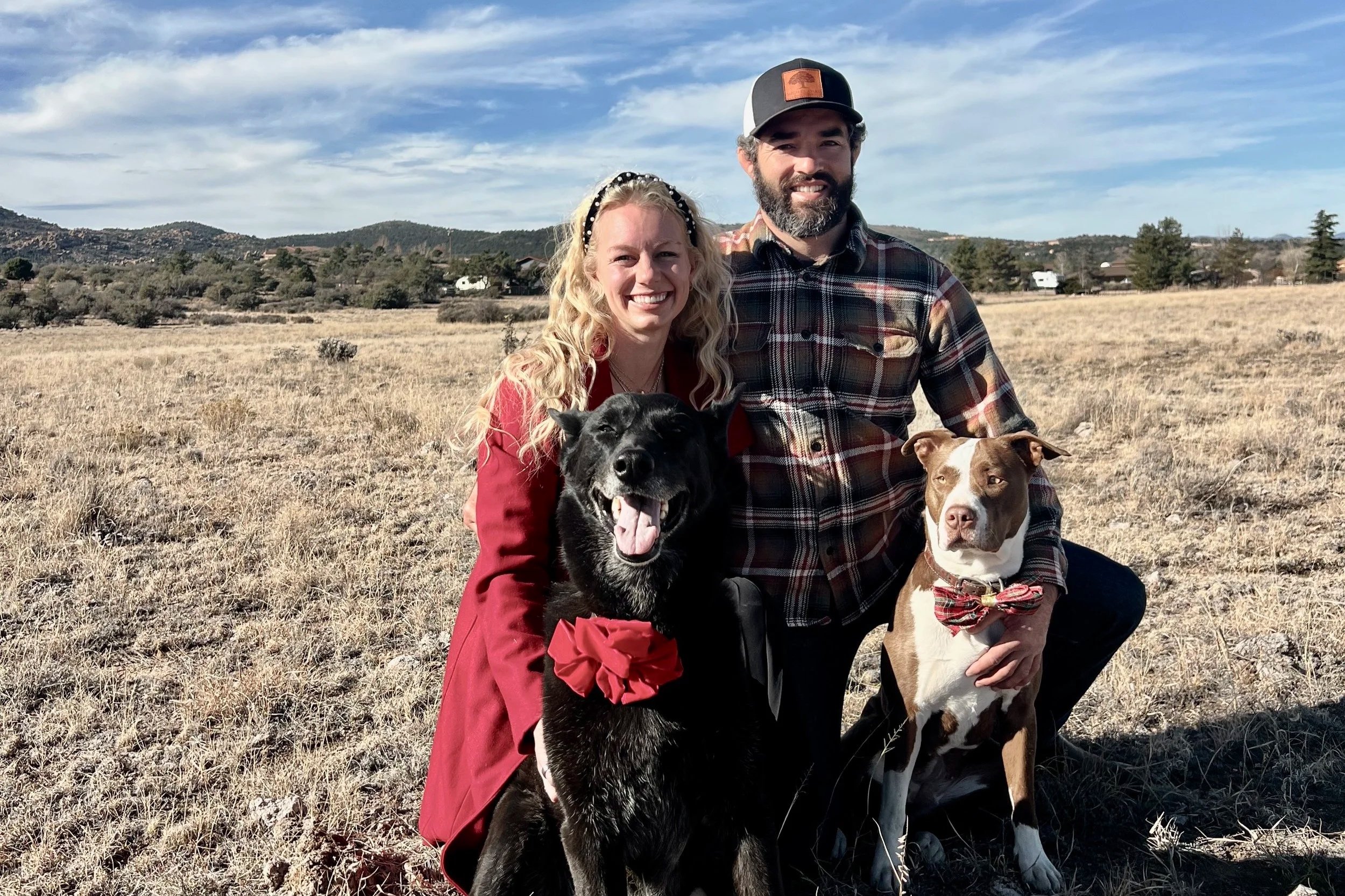 A smiling man and woman kneeling outdoors in a field with two dogs, one black and one brown and white, under a partly cloudy sky