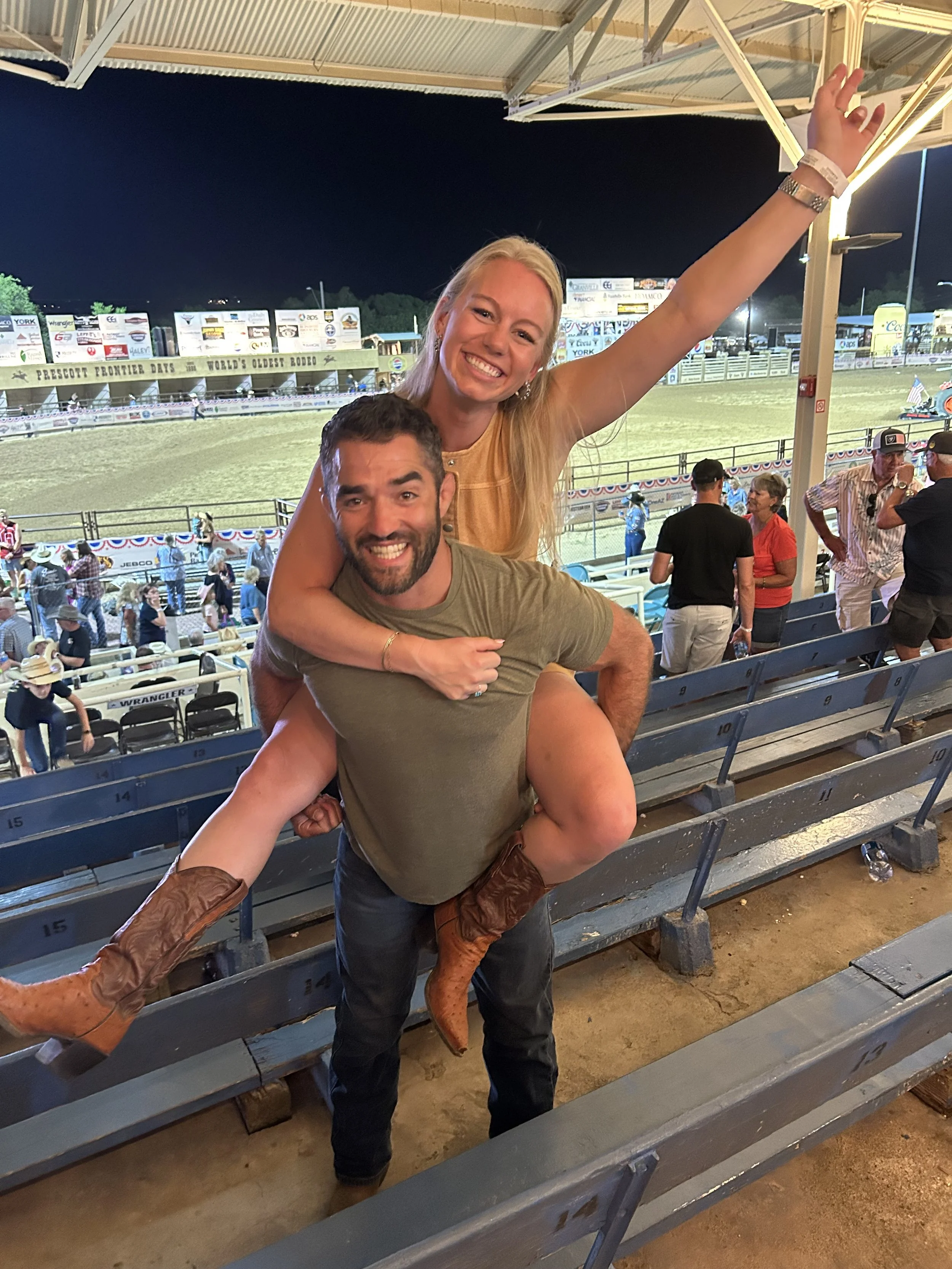 A man giving a piggyback ride to a woman at a rodeo arena during nighttime, both smiling and posing for the camera.