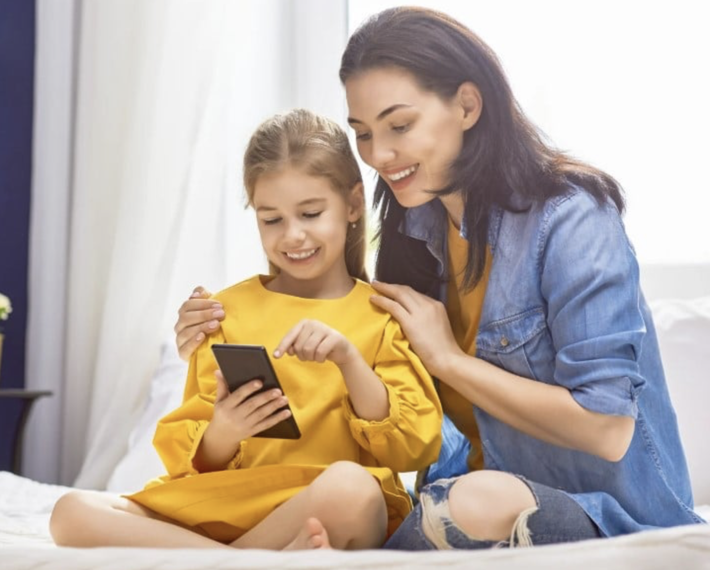 A woman and girl sitting on a bed looking at a smartphone together, smiling.
