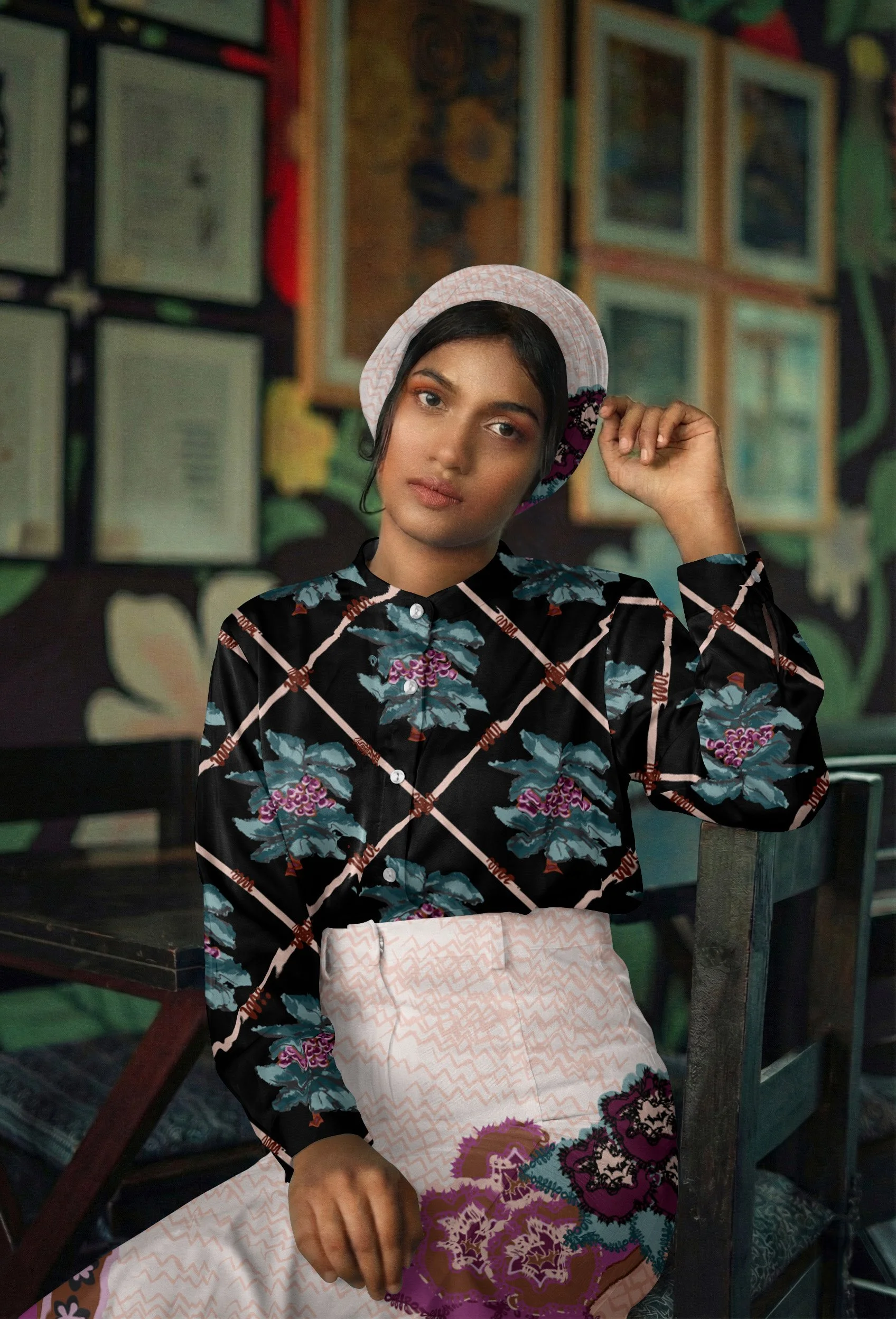 A woman wearing a floral-patterned blouse, a pink hat, and a patterned skirt, seated in an art gallery surrounded by framed artwork and colorful murals.