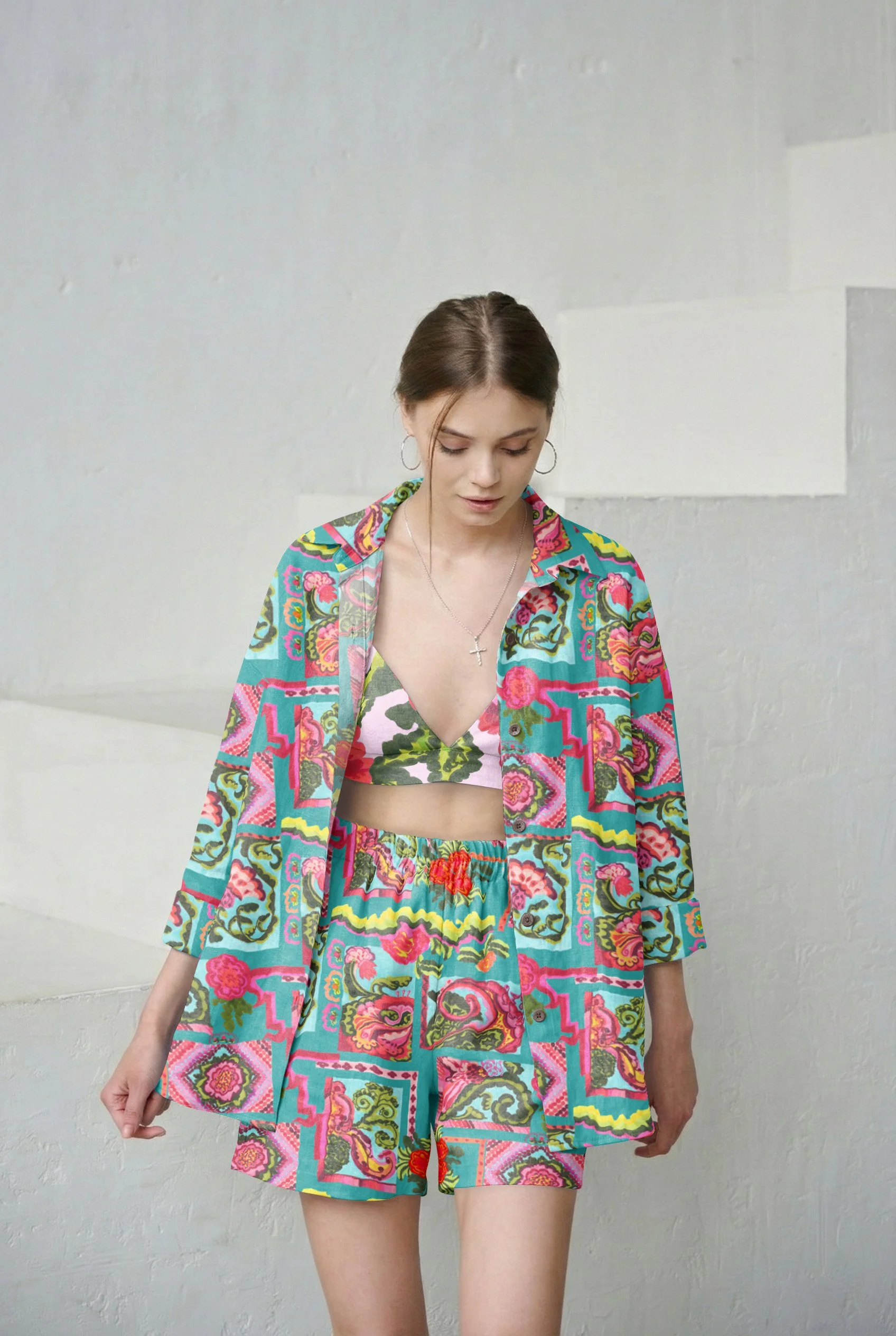 A young woman with brown hair, wearing bohemian-style clothing with a floral and geometric pattern in bright colors, standing against a plain, light-colored wall.