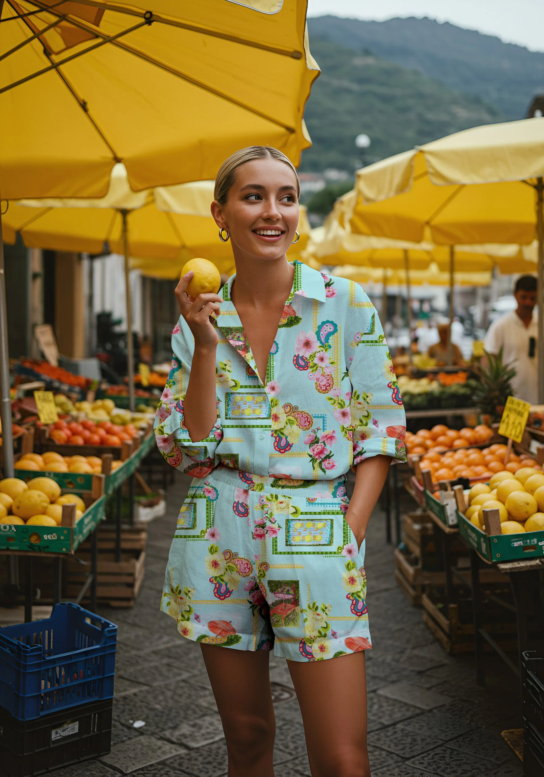 A young woman with blonde hair smiling, holding a lemon, at an outdoor fruit market with yellow umbrellas and stalls of fresh produce.